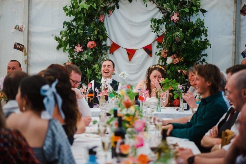 a bride and groom laughing at the end of a wedding table with their guests in a marquee 