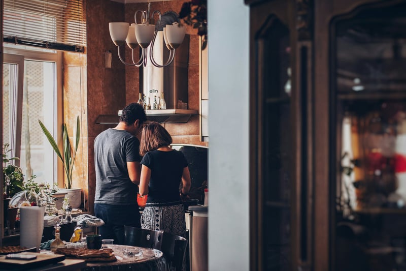 Un couple cuisine ensemble dans une cuisine chaleureuse et décorée, vu de l’arrière, entouré de plantes et d’objets du quotidien.