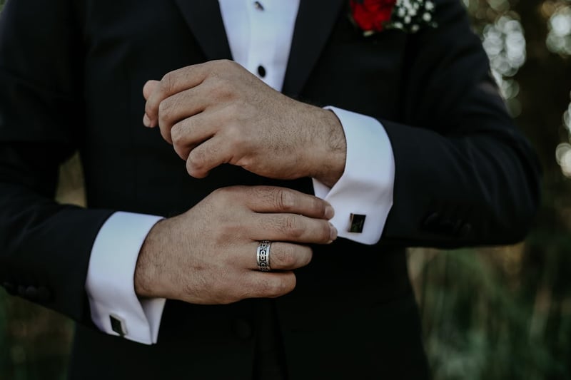 Gros plan sur un marié en costume noir ajustant ses boutons de manchette argentés, avec une bague élégante et une boutonnière rouge en arrière-plan.