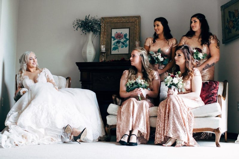 Une mariée détendue et souriante en robe blanche en dentelle, assise sur un canapé, entourée de ses demoiselles d’honneur vêtues de robes dorées scintillantes, tenant des bouquets de fleurs.