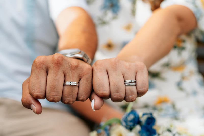 Deux mains entrelacées montrant leurs alliances, avec un arrière-plan flou de fleurs et de vêtements colorés.