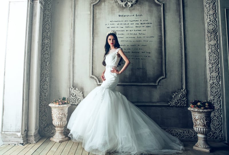 Une mariée dans une robe élégante et un long voile en tulle flottant dans le vent, avec un fond de verdure et un ciel couvert.