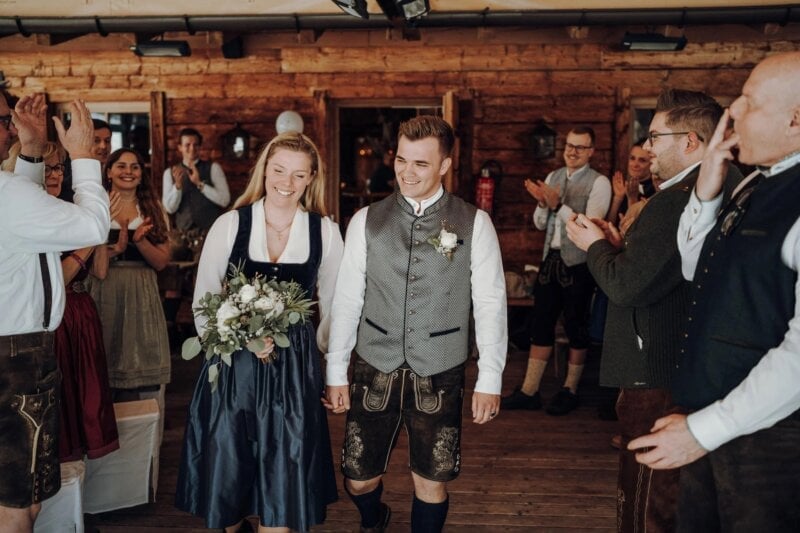 Brautpaar in Tracht und Dirndl wird von applaudierenden Gästen in Holzhütte empfangen