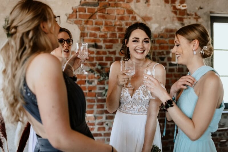 Braut stößt lachend mit Freundinnen an, Glückwünsche zur Hochzeit vor Backsteinwand