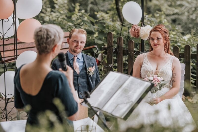 Traurednerin liest Sprüche zur Hochzeit vor, Brautpaar sitzt lächelnd davor