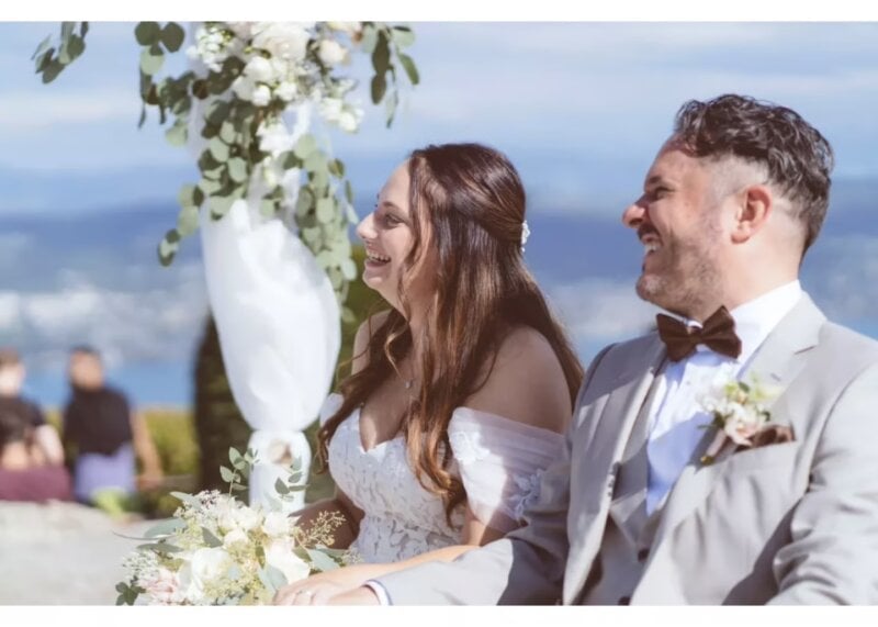 Lachendes Brautpaar bei der Trauung mit Blumenbogen und Meerblick im Hintergrund