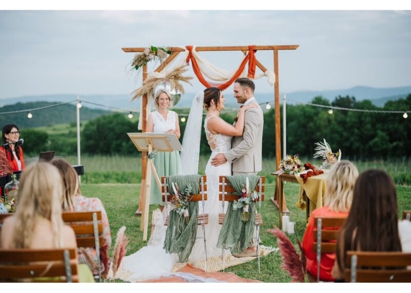 Rednerin spricht Glückwünsche zur Hochzeit, Brautpaar umarmt sich vor Traubogen