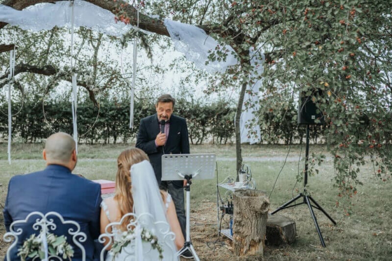 Vater des Bräutigams spricht Hochzeitsrede am Mikrofon unter blühendem Baum