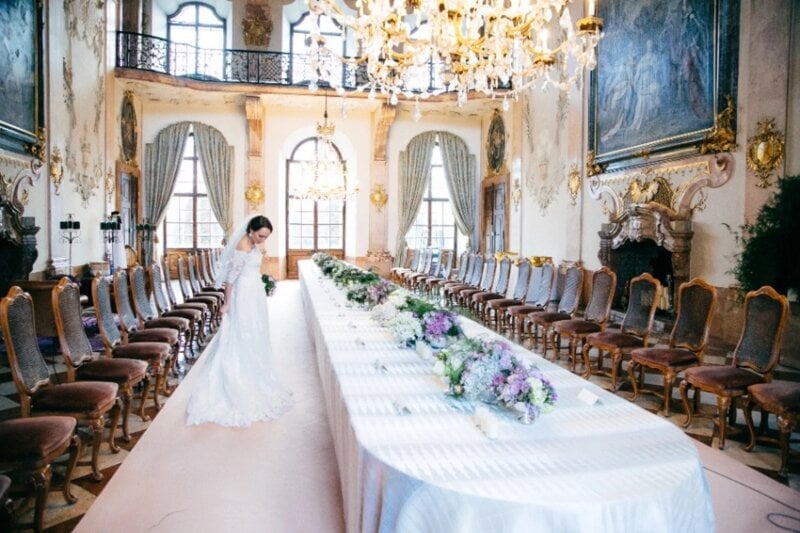 Braut steht allein in prunkvollem Schlosssaal mit Kronleuchter und festlich gedeckter Tafel