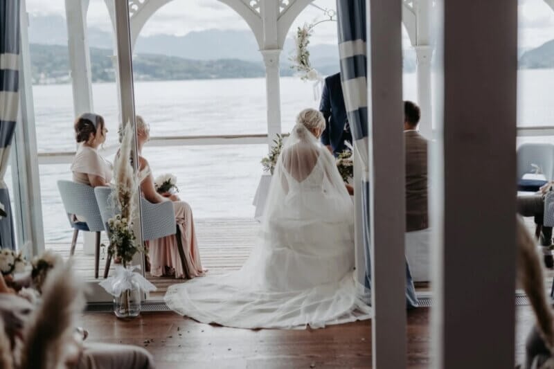 Braut mit langer Schleppe steht bei der Trauung in einem Pavillon am See