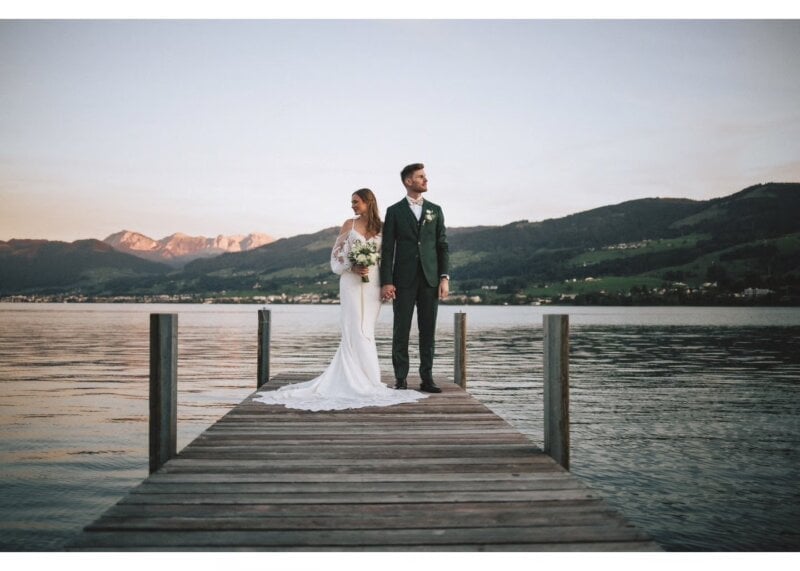 Brautpaar steht auf einem Bootssteg am See mit Bergpanorama im Abendlicht