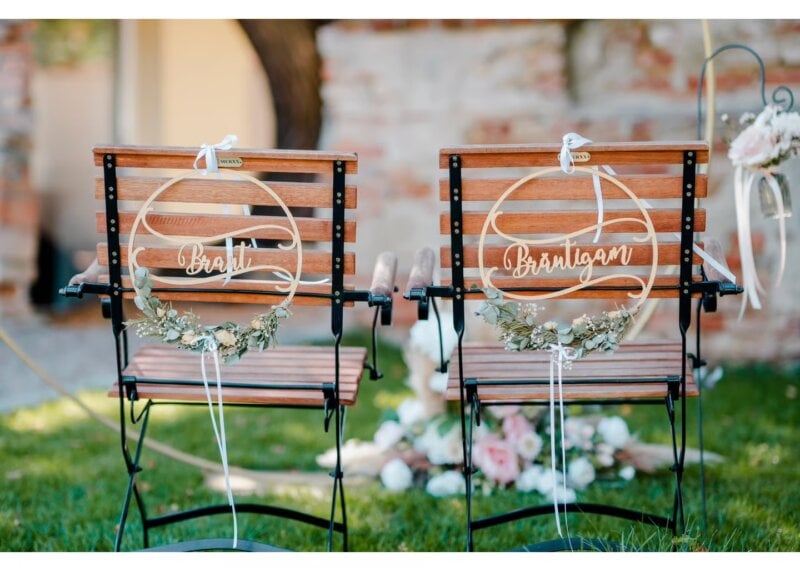 Zwei dekorierte Holzstühle mit Schildern „Braut“ und „Bräutigam“ und Blumenschmuck im Garten