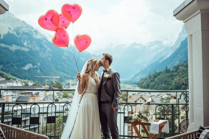 Bräutigam küsst Braut auf die Stirn, sie hält herzförmige Luftballons vor Bergpanorama