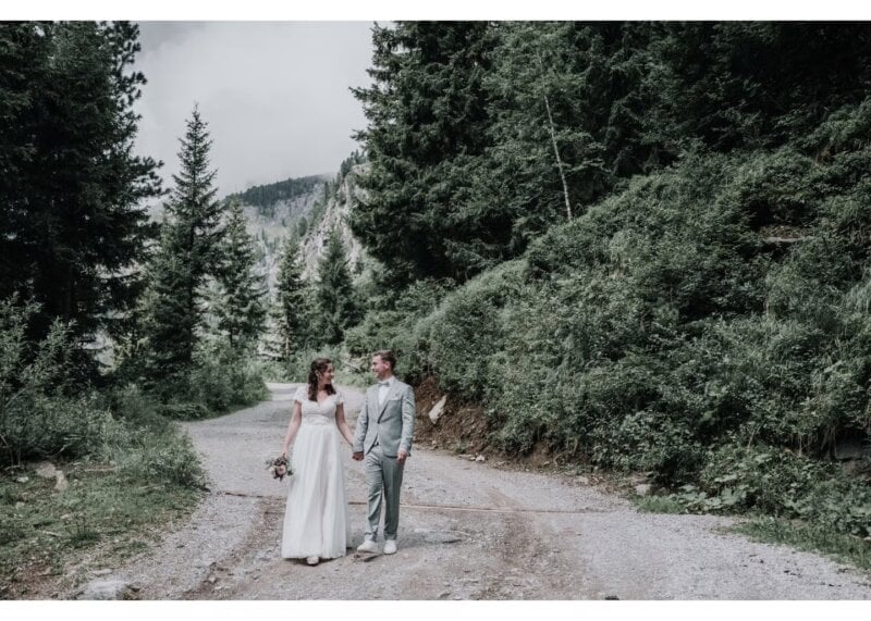 Brautpaar geht Hand in Hand auf einem Waldweg umgeben von Bergen und Tannen