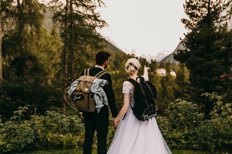 Brautpaar mit „Just Married“-Rucksäcken wandert Hand in Hand durch einen Bergwald