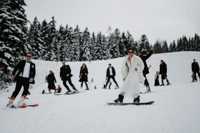 Brautpaar und Hochzeitsgäste fahren im Brautkleid und Anzug Ski und Snowboard auf verschneiter Piste