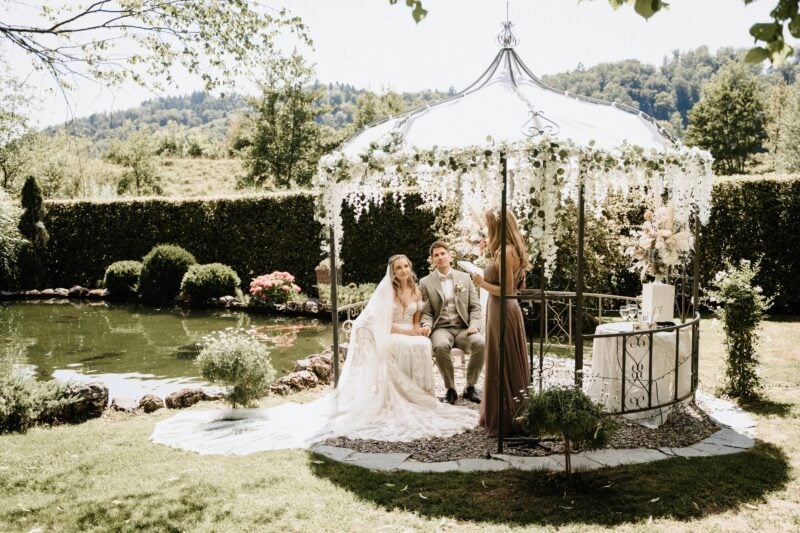 Brautpaar sitzt bei der Trauzeremonie in blumengeschmücktem Pavillon am idyllischen Teich im Garten
