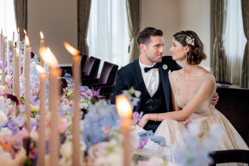 Brautpaar sitzt Hand in Hand an festlich mit Kerzen und Blumen dekorierter Tafel