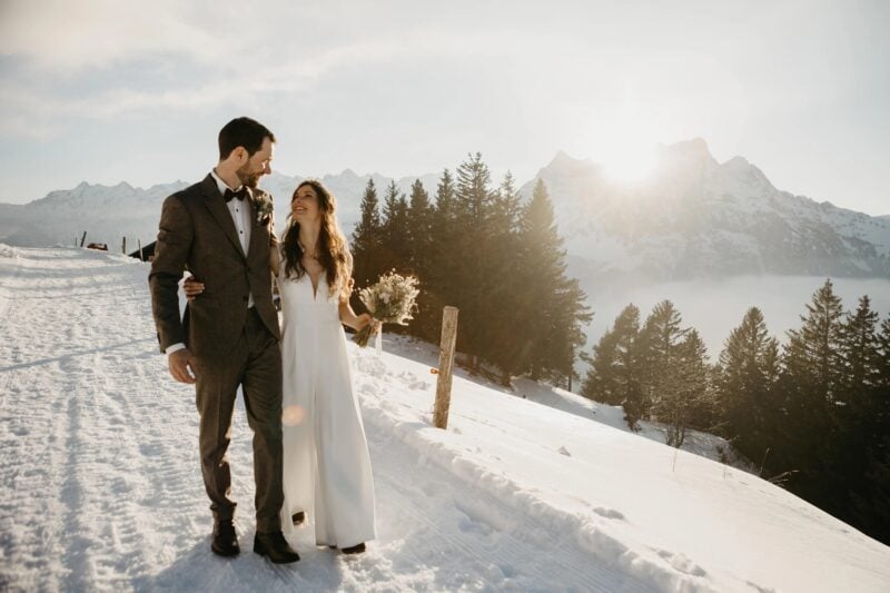 Brautpaar geht Arm in Arm im Schnee vor sonnigem Bergpanorama mit Wolkenmeer