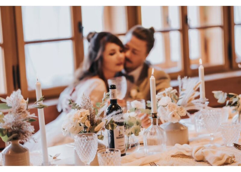 Brautpaar kuschelt im Hintergrund hinter festlich gedeckter Tafel mit Kerzen, Blumen und Weinflasche vom Weingut