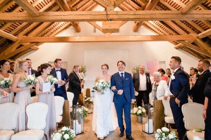 Vater führt Braut mit Blumenstrauß durch den Mittelgang der Trauung in Scheune mit Holzbalken