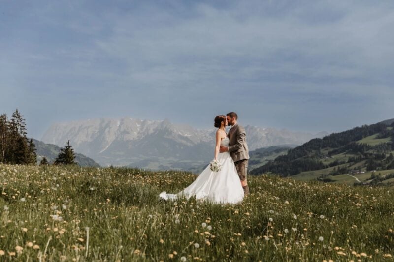 Brautpaar küsst sich auf Blumenwiese vor Alpenpanorama