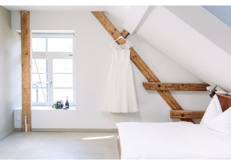 Brautkleid hängt an Holzbalken im Dachzimmer neben Brautstrauß und Champagner