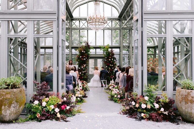 Trauung in einem lichtdurchfluteten Glaspavillon mit üppiger Blumendekoration und Kronleuchter