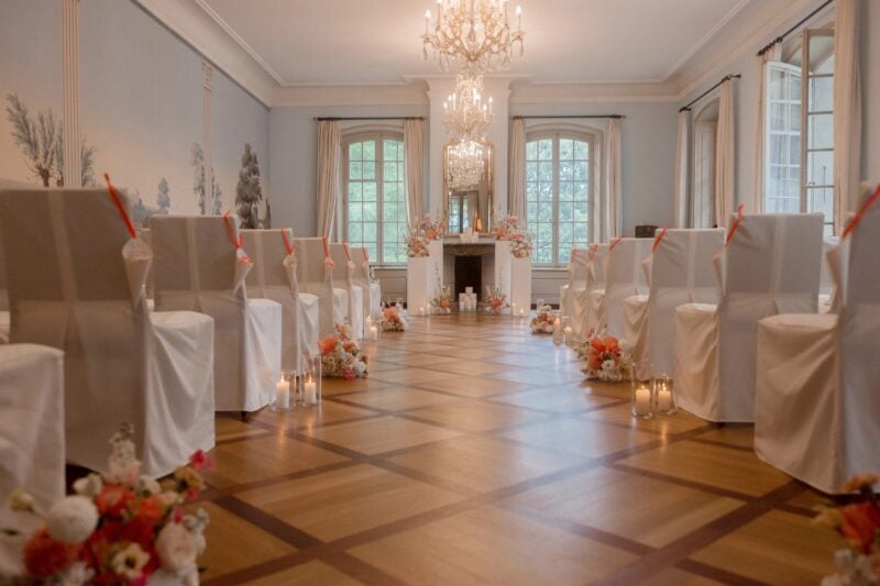 Festlich dekorierter Trausaal in Schloss Hünigen mit weissen Hochzeitsstühlen und Blumendeko in Koralle und Rosa