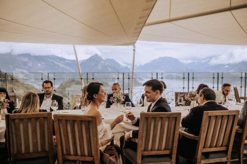 Brautpaar stößt lächelnd mit Wein an der festlichen Tafel unter einem Sonnensegel mit Bergpanorama an