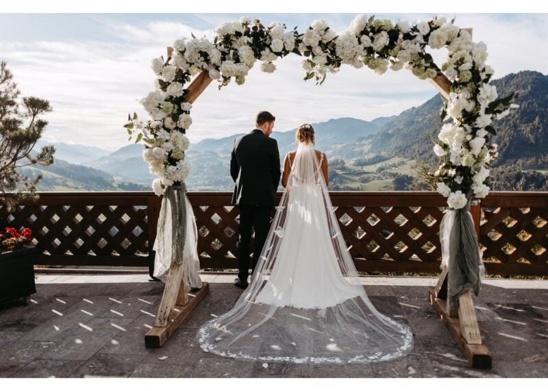 Brautpaar steht Hand in Hand unter einem Blumenbogen mit Blick auf ein Bergpanorama