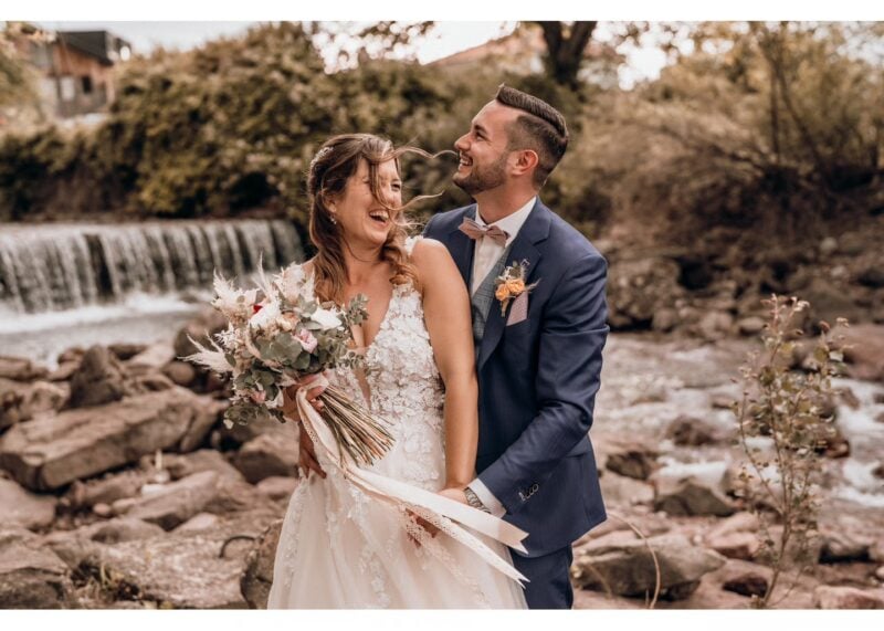Brautpaar am Fluss mit Boho-Brautstrauss aus Trockenblumen, Bräutigam im marineblauen Anzug mit rosa Fliege