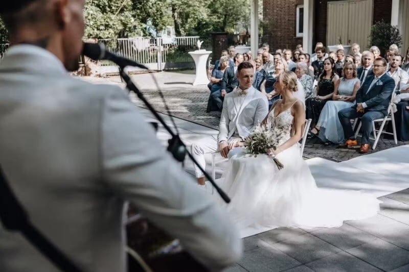 Hochzeitssänger mit Mikrofon singt vor sitzendem Brautpaar und Gästen bei Trauung im Freien