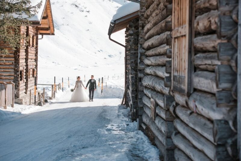 Brautpaar geht Hand in Hand durch ein verschneites Bergdorf zwischen alten Holzhütten