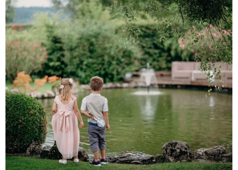 Kleine Blumenkinder in festlicher Kleidung schauen auf Teich mit Springbrunnen im Garten