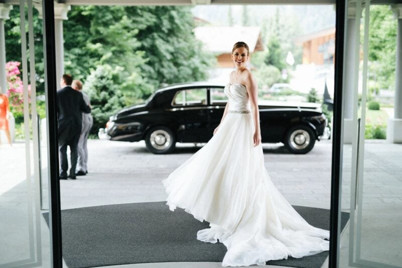 Braut in fließendem Hochzeitskleid steht am Hoteleingang vor einem klassischen schwarzen Oldtimer