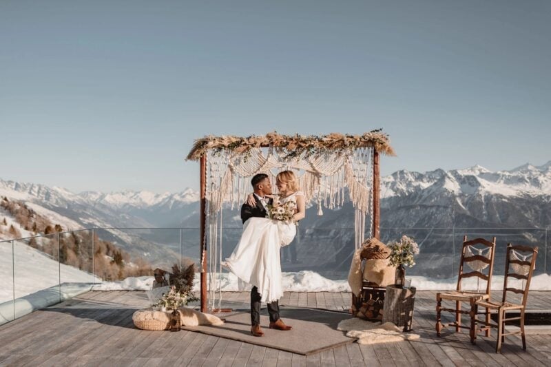 Bräutigam trägt Braut auf Armen vor Boho-Traualtar mit Alpenpanorama