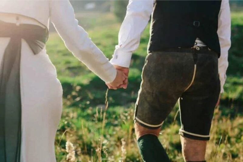 Brautpaar hält Hände, Bräutigam trägt traditionelle Lederhose zur Hochzeit