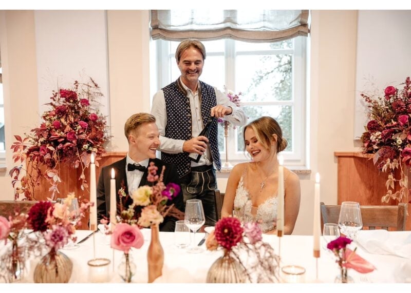 Brautpaar sitzt lachend an festlich dekorierter Hochzeitstafel, während ein Kellner Wein einschenkt