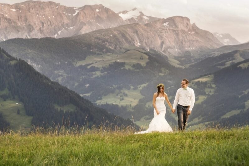Brautpaar geht Hand in Hand über eine grüne Bergwiese vor eindrucksvollem Alpenpanorama