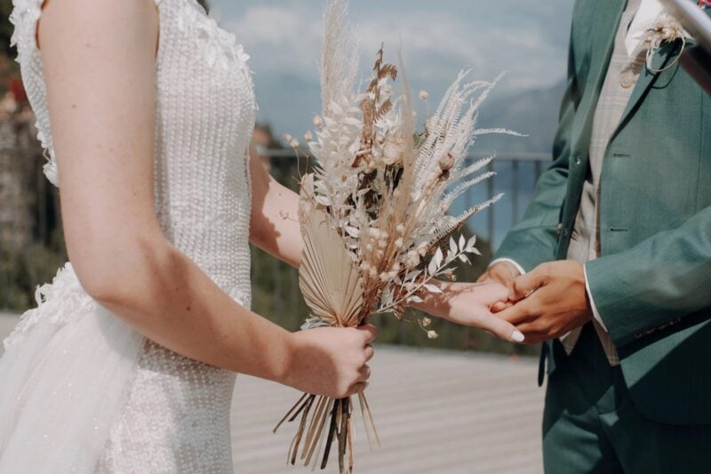 Brautpaar hält Hände mit getrocknetem Bouquet, Bräutigam im salbeigrünen Anzug – harmonisch abgestimmte Hochzeitsfarben