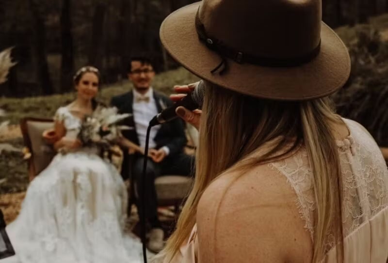 Hochzeitssängerin mit Hut singt vor sitzendem Brautpaar bei Waldtrauung