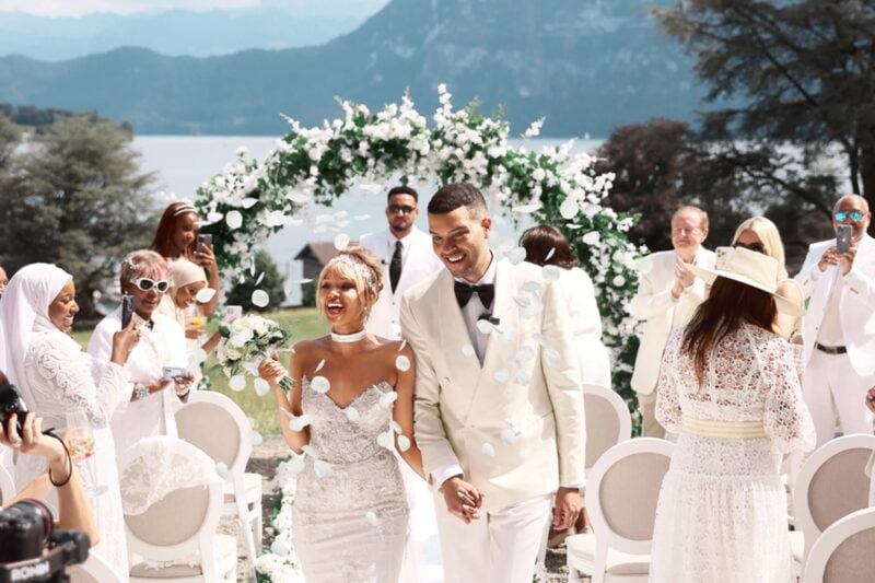 Brautpaar geht lachend durch Konfettiregen zwischen Gästen bei einer Seehochzeit vor Bergpanorama