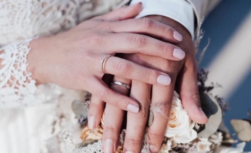 Hände eines Paares mit Verlobungs- und Eheringen über dem Brautstrauß – Hochzeit im Das Teichwerk