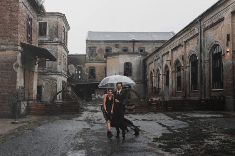 Brautpaar unter einem transparenten Schirm im Regen im Industrie-Innenhof der Papierfabrik Varieté – urbaner Stil der Hochzeitsfotografie