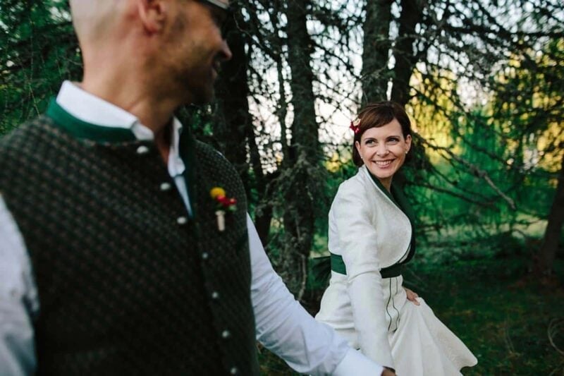 Bräutigam in Trachtenweste führt die Braut an der Hand durch den Wald – Hochzeit in Tracht