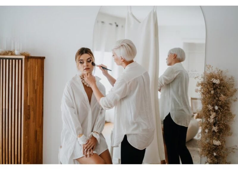 Braut bekommt vor dem Spiegel Puder-Make-up von ihrer Visagistin aufgetragen