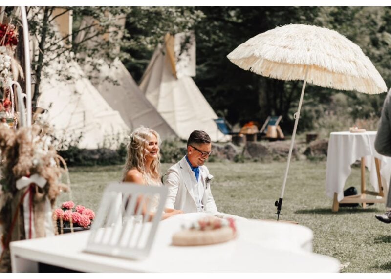 Brautpaar sitzt entspannt an der Festtafel vor Tipis im Boho-Stil – perfektes Timing für die Hochzeitsunterhaltung