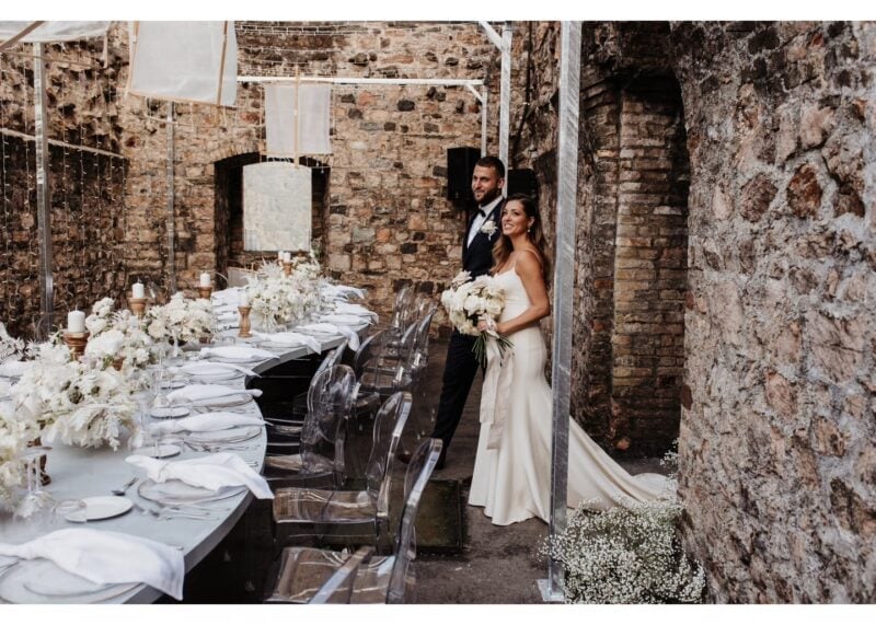 Brautpaar neben einer festlich gedeckten Hochzeitstafel mit weissen Blumen zwischen alten Steinmauern der Tenuta Castello di Morcote