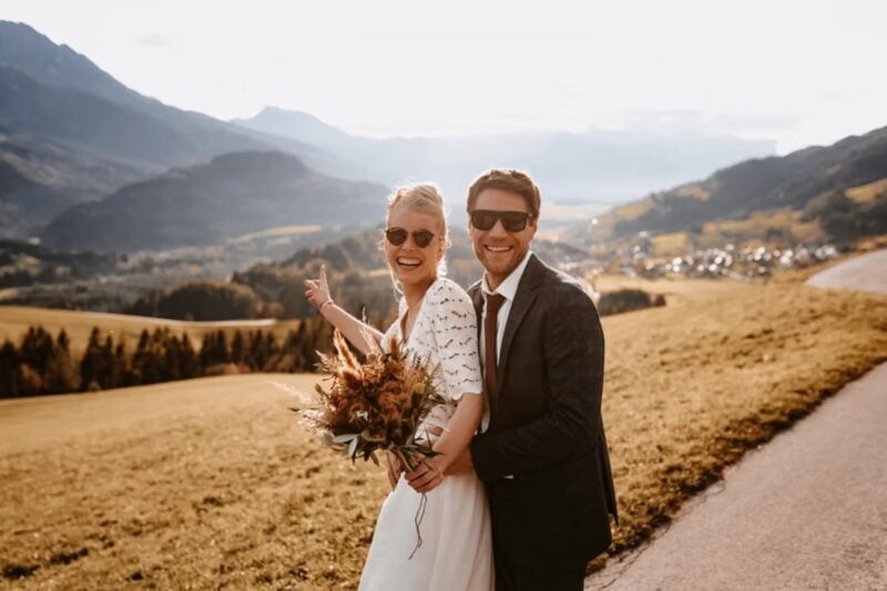 Brautpaar mit Sonnenbrille lacht ausgelassen vor Bergpanorama